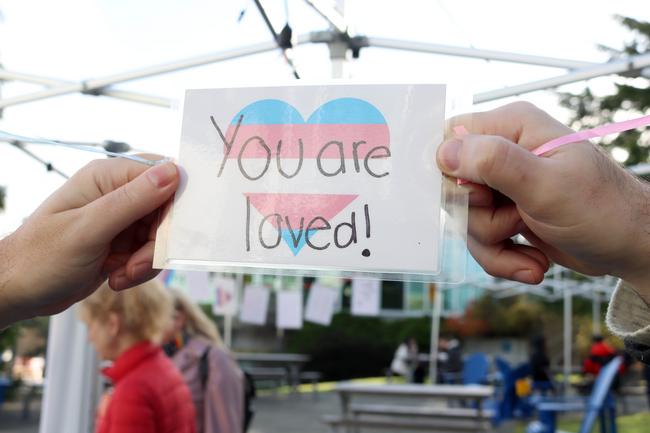Two hands hold up a sign that says you are loved with a heart with the transgender flag on it
